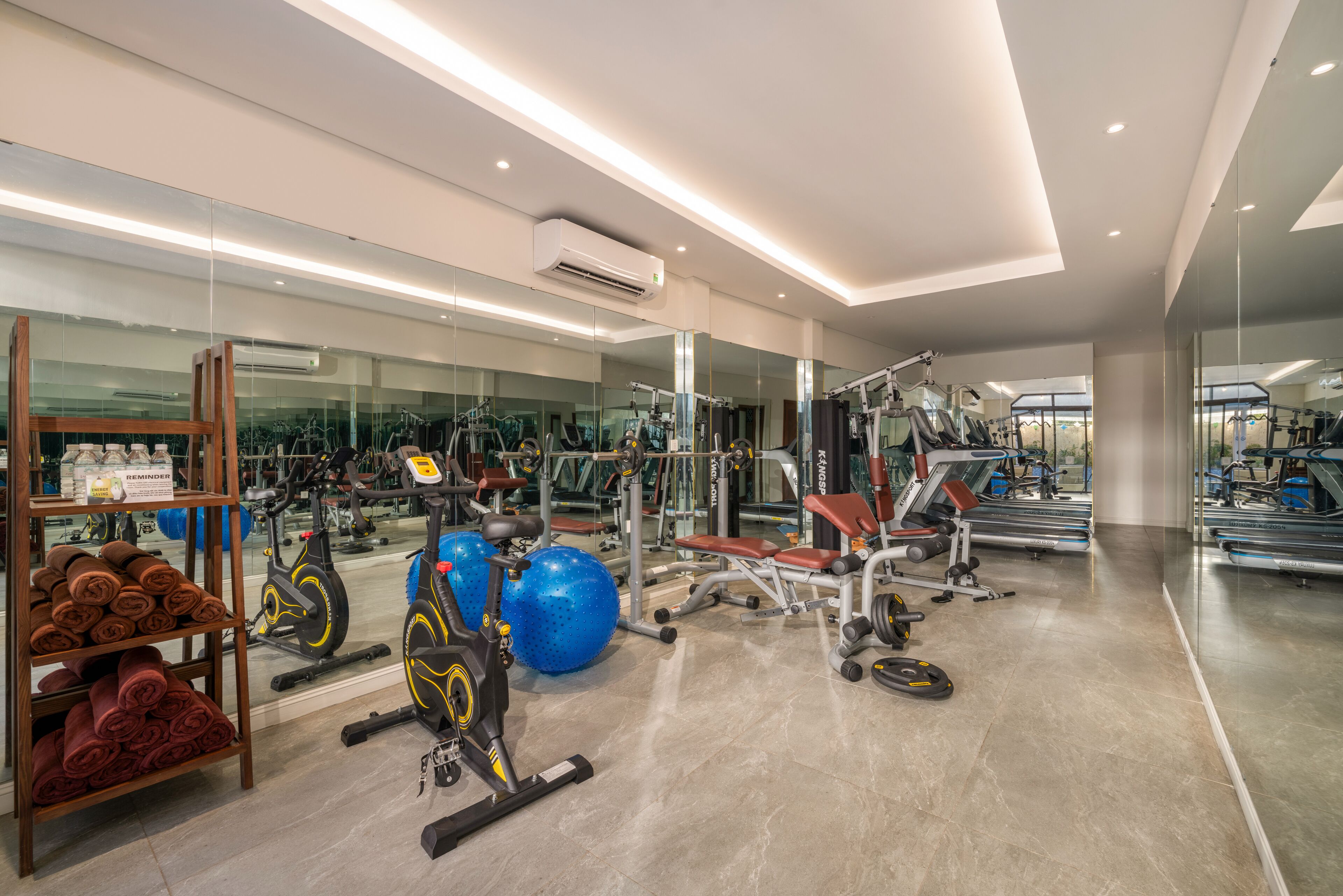 A modern gym with various exercise equipment, a mirrored wall, and a wooden rack with towels.