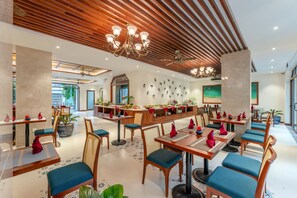 Un vaste espace dédié à la salle à manger, avec un plafond orné de panneaux en bois, de nombreuses tables entourées de chaises rembourrées bleues, et un buffet central.