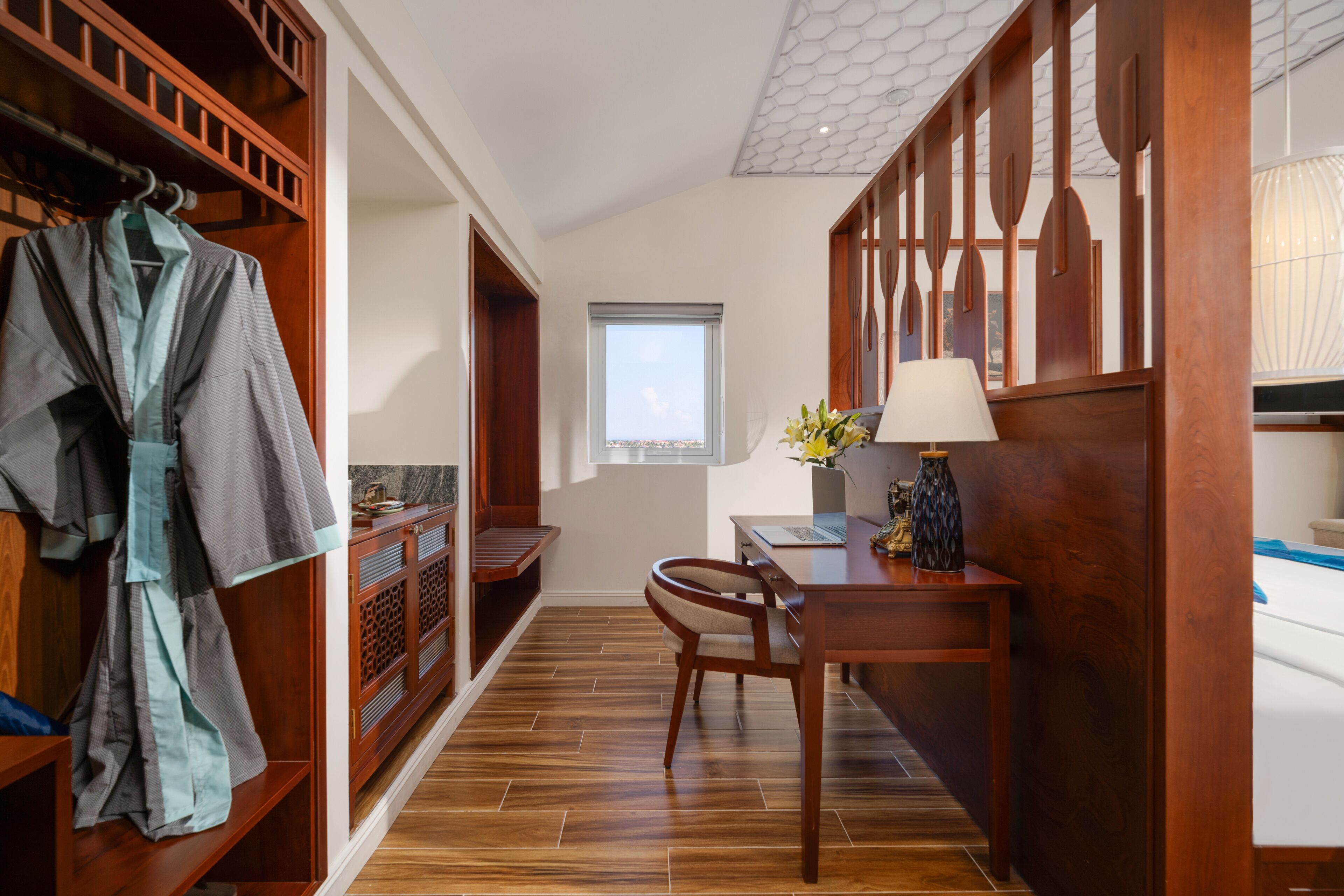 A hotel room with a wooden floor, a desk with a lamp, a chair, and a robe hanging on a rack.