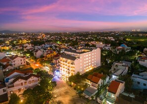 Une vue aérienne d’une ville au crépuscule, avec un hôtel imposant à plusieurs étages et doté de nombreux balcons, entouré d’immeubles résidentiels et bordé d’une rue bien éclairée.