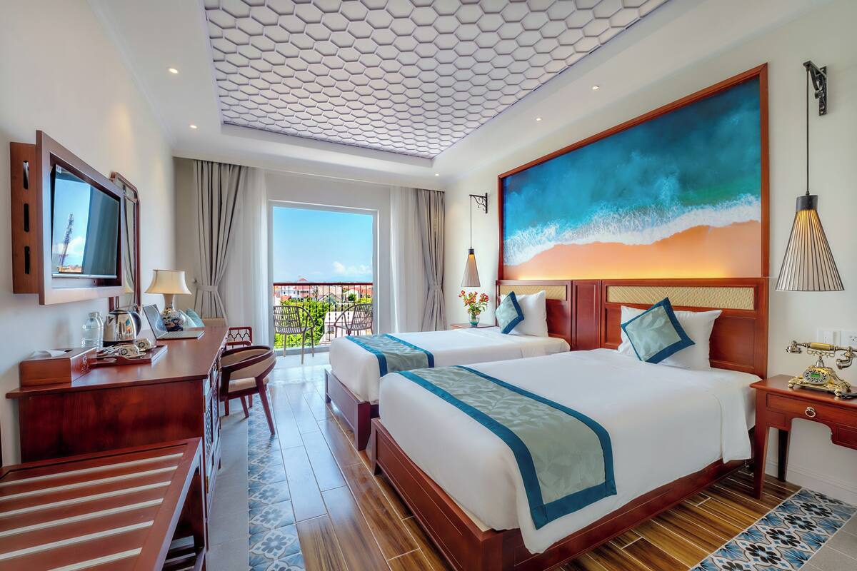 Hotelkamer met twee bedden, een bureau, een televisie, een balkon met uitzicht en een groot schilderij van een strand.