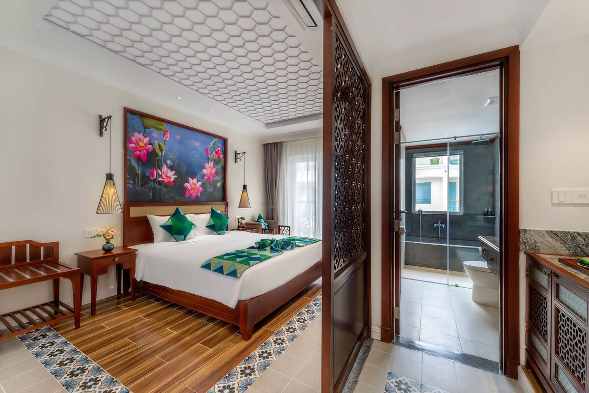 Een hotelkamer met een groot bed, een bloemen schilderij, een houten bankje, een nachtkastje en uitzicht op het balkon en de badkamer.