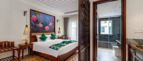 Habitación de hotel con una cama grande, un cuadro de flores, un banco de madera, una mesita de noche y vistas al balcón y al baño.