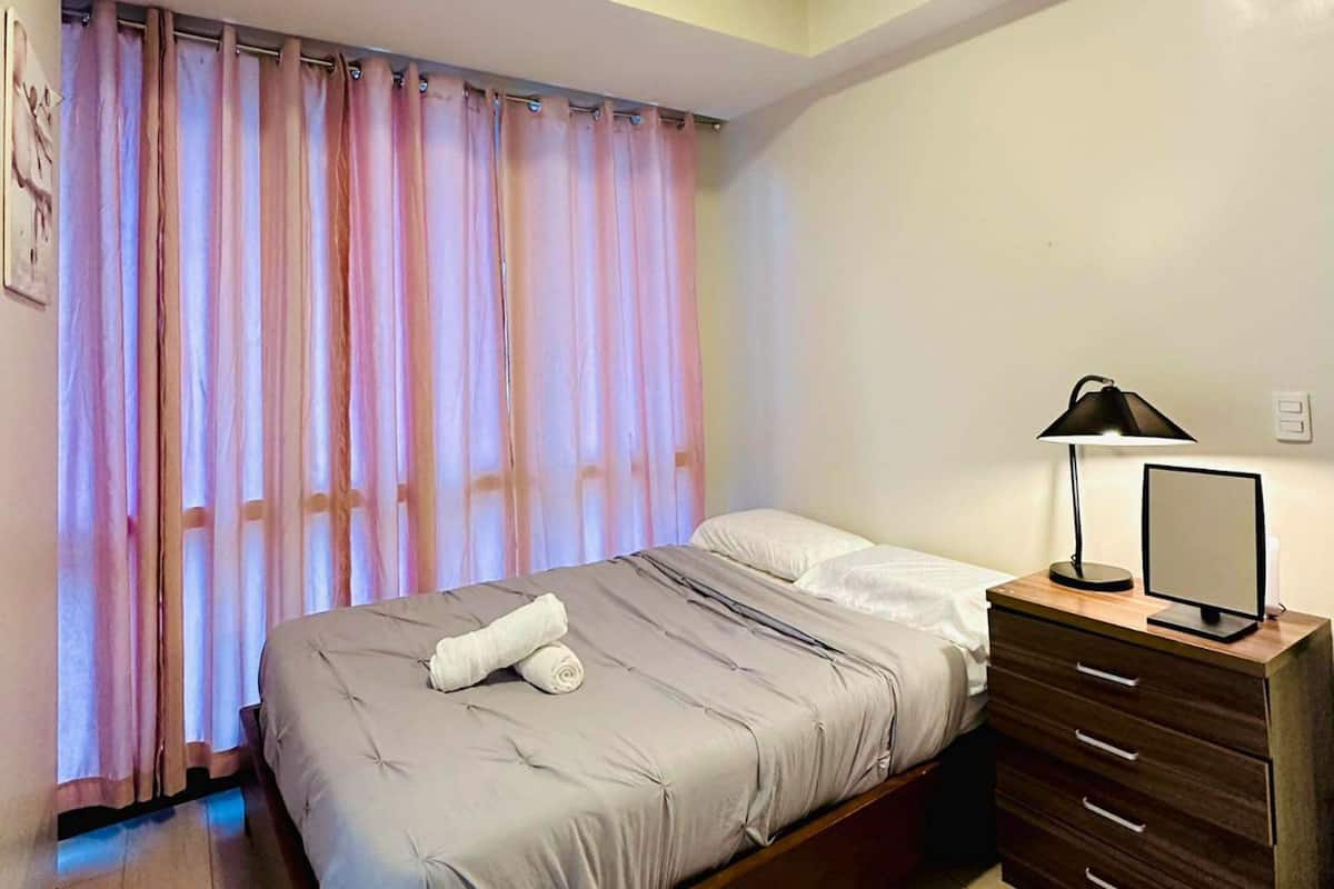 A bedroom with a bed, a bedside table with a lamp, a mirror, and a window with curtains.
