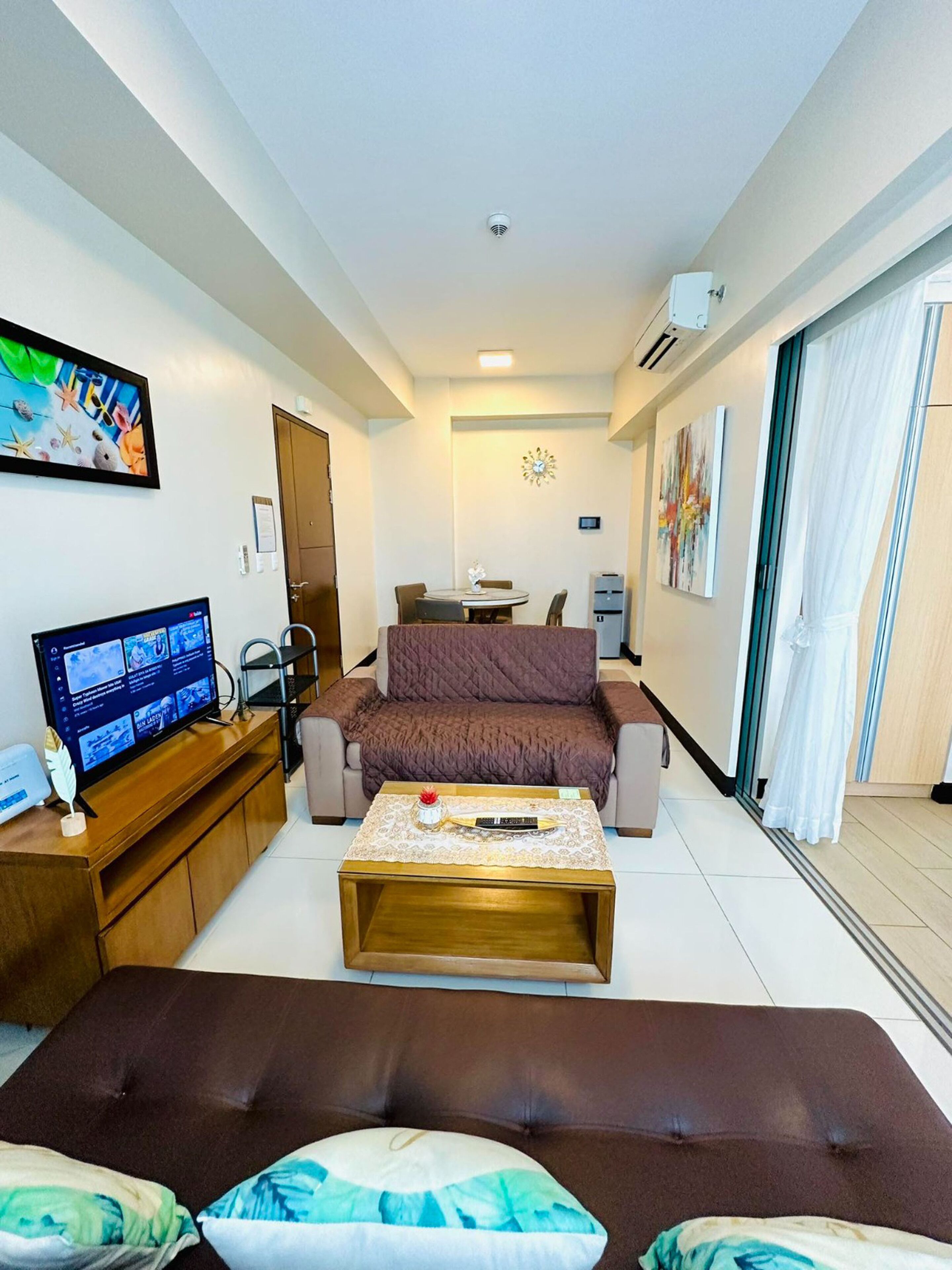 Una sala de estar con un sofá marrón, una mesa de centro, un televisor sobre un mueble de madera y un cuadro en la pared.