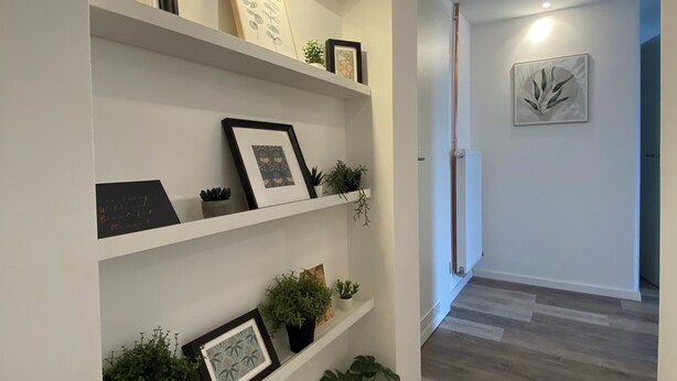 A hallway with white walls, wooden flooring, and built-in shelving with framed pictures and potted plants.