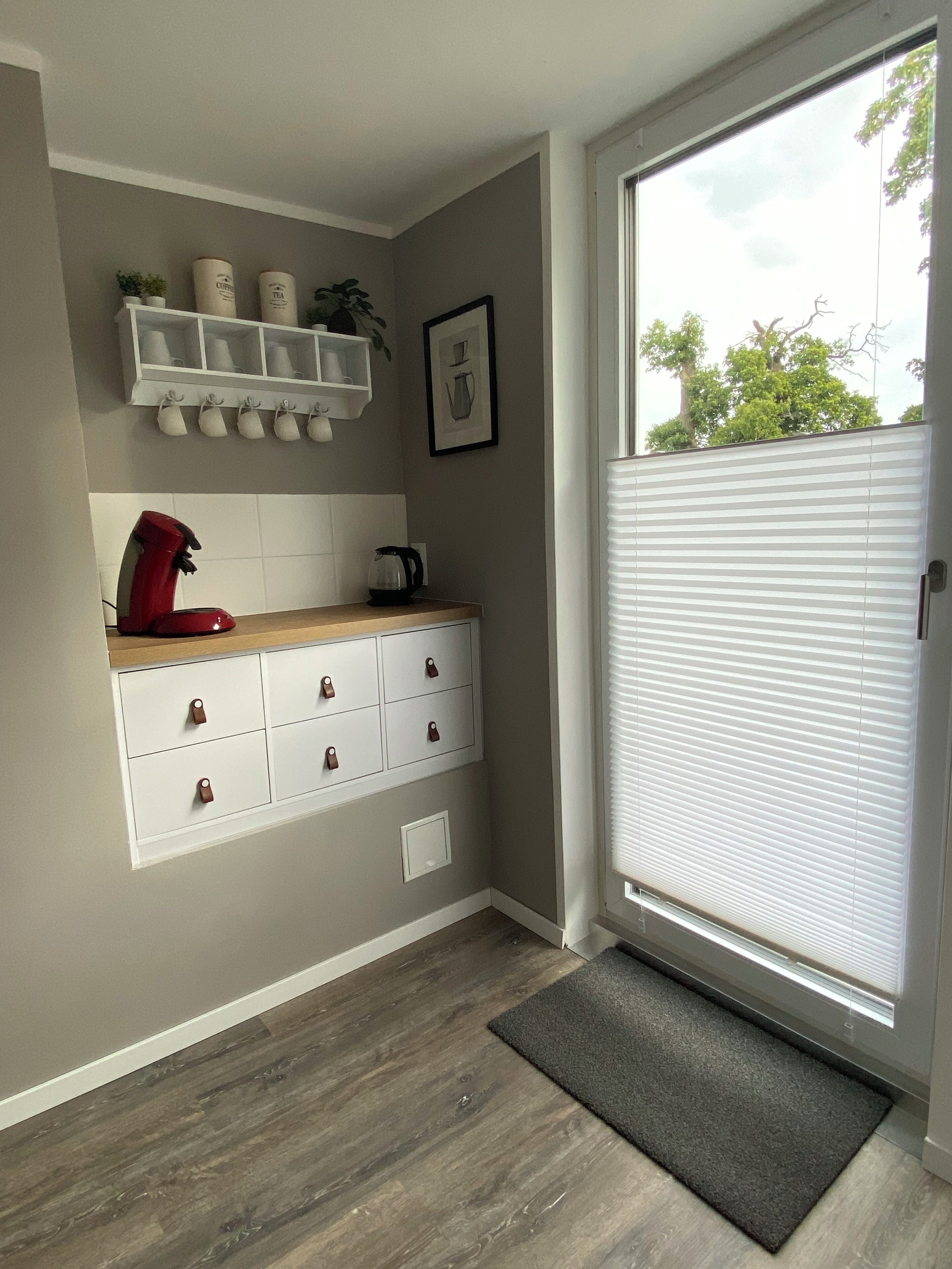 A modern kitchen with a built-in coffee machine, a white cabinet with drawers, a countertop, and a window with blinds.