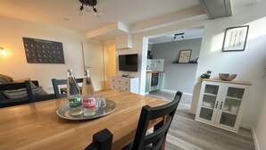 A modern dining area with a wooden table, black chairs, and a tray with bottles. A kitchen is visible in the background with a TV and cabinets.