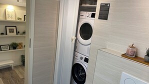 A laundry room with stacked washing machines, a bench, and a shelf with plants and framed pictures.
