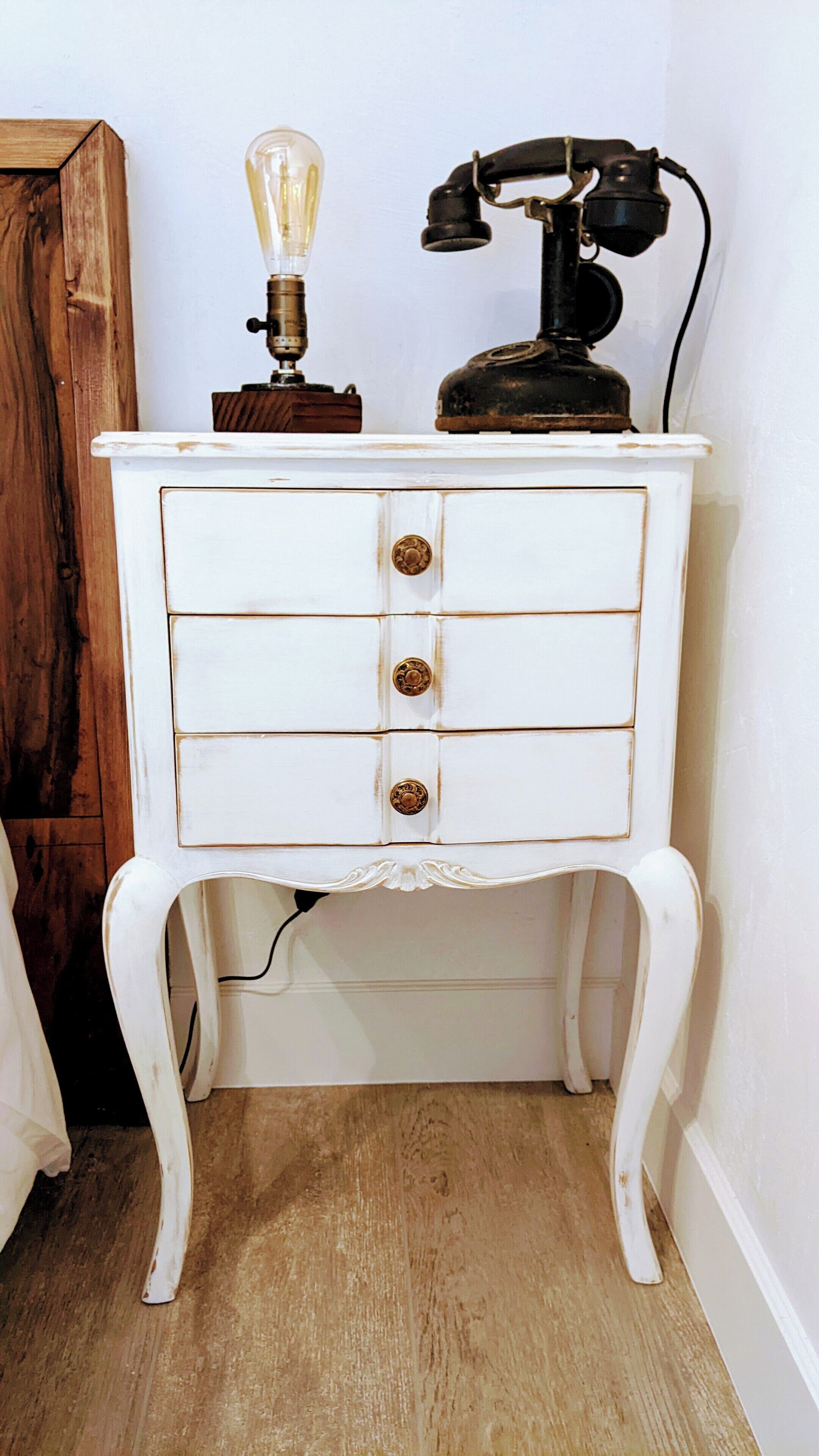 Un aparador blanco de estilo antiguo con tres cajones y pomos de latón, un teléfono antiguo y una bombilla en la parte superior.