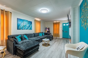 A living room with a sectional sofa, a small round coffee table, a white armchair, and a flat-screen TV mounted on the wall.
