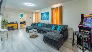 Uma sala de estar com sofá modular, mesa de centro, relógio, quadro na parede e console de jogos.