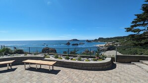 Un pati amb una cadira de fusta i una jardineria de pedra amb flors, amb vista a una zona costanera amb roques i una platja.