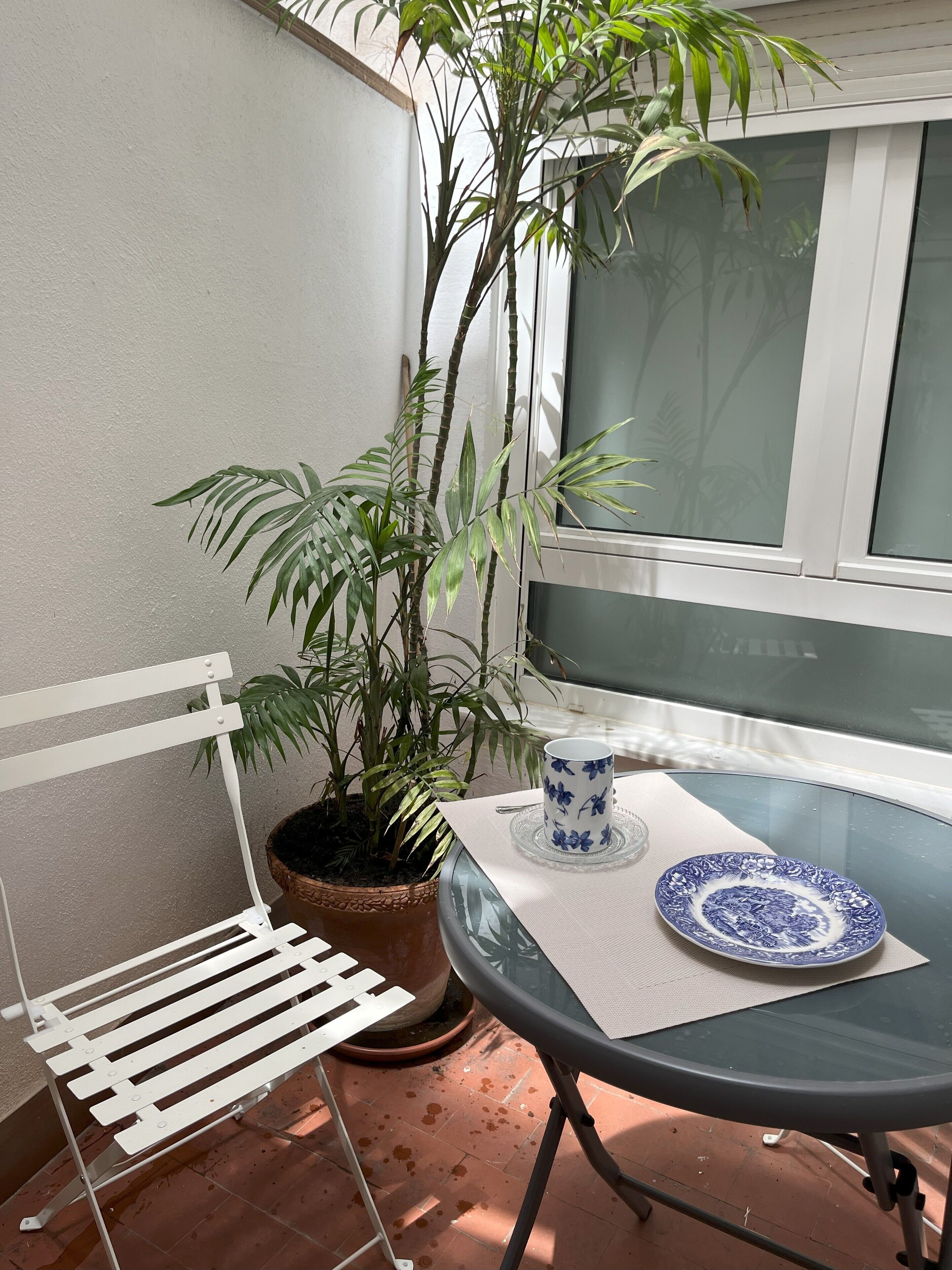 Eine kleine Terrasse mit einem weißen Metallstuhl, einem Glastisch, einem Blumentopf und einem Fenster.