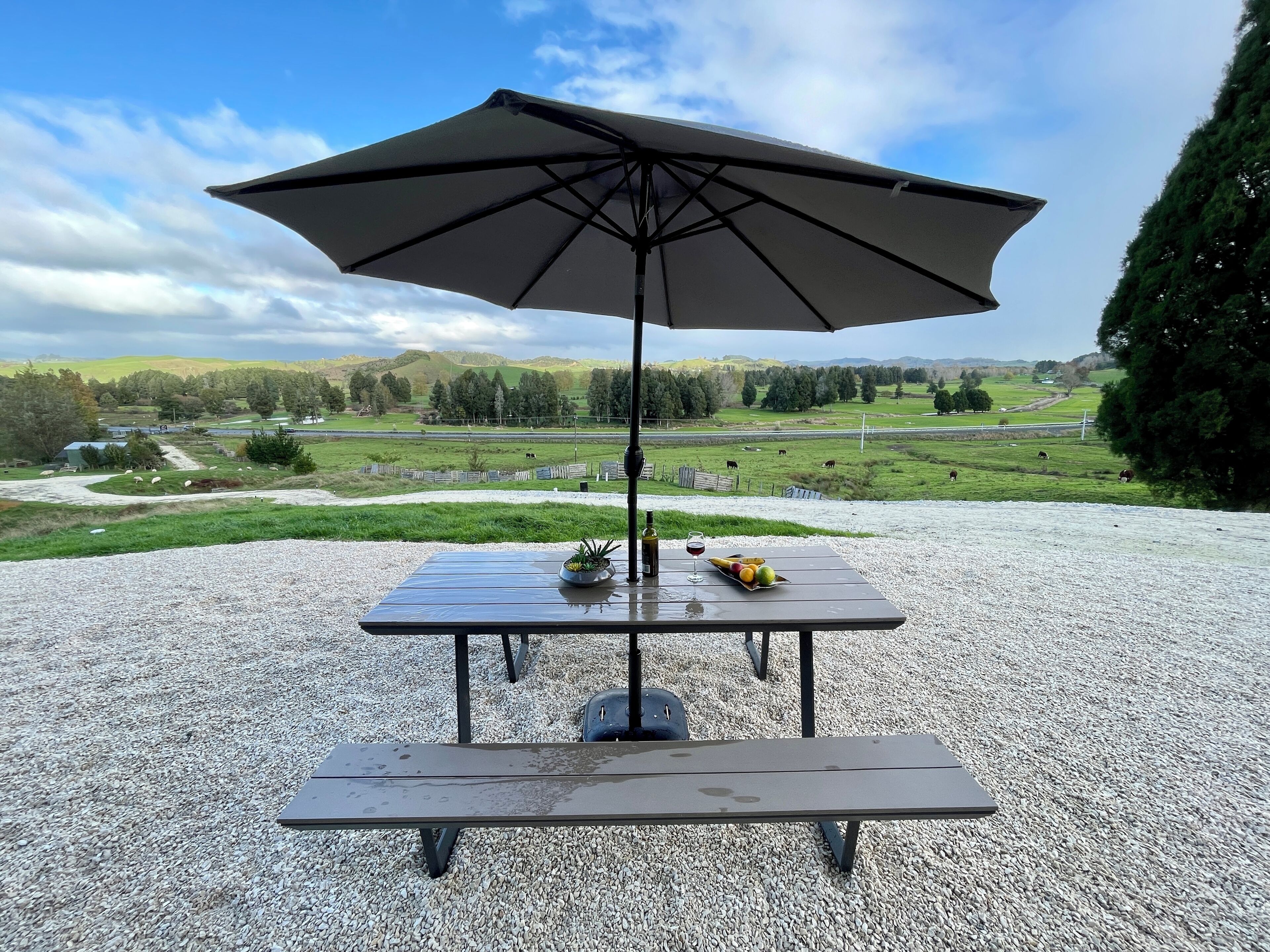Una mesa con sombrilla, una botella y fruta sobre un suelo de grava con una vista panorámica al fondo.