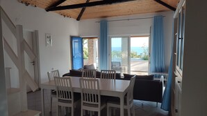 A dining area with a wooden ceiling, white chairs, a table, and a view of the sea through a door.