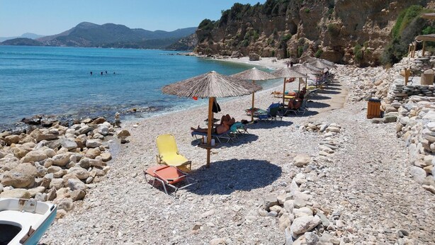 A pebble beach with umbrellas and sun loungers, a rocky shoreline, and mountains in the background.