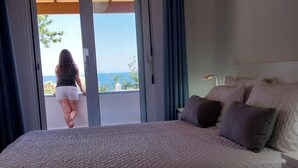 A bedroom with a large bed, a view of the sea through a sliding door, and a nightstand with a lamp.