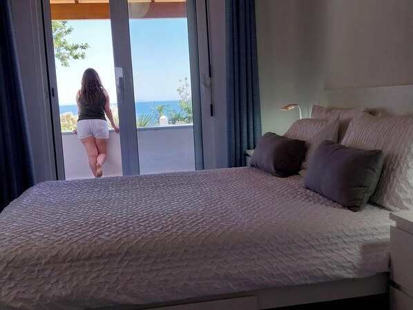 A bedroom with a large bed, a view of the sea through a sliding door, and a nightstand with a lamp.