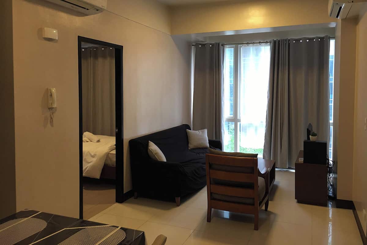 Hotelkamer met een bed, een zwarte bank, een houten stoel, een kleine tafel en een bureau met een computer. Er is een raam met gordijnen, een airconditioning en een telefoon aan de muur.