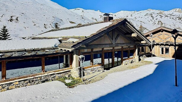 A wooden chalet with large windows, a stone foundation, and a snow-covered mountain backdrop.