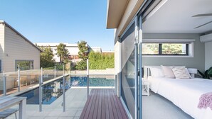 Une chambre moderne avec un grand lit, un balcon vitré donnant sur une piscine et une vue sur les maisons voisines.