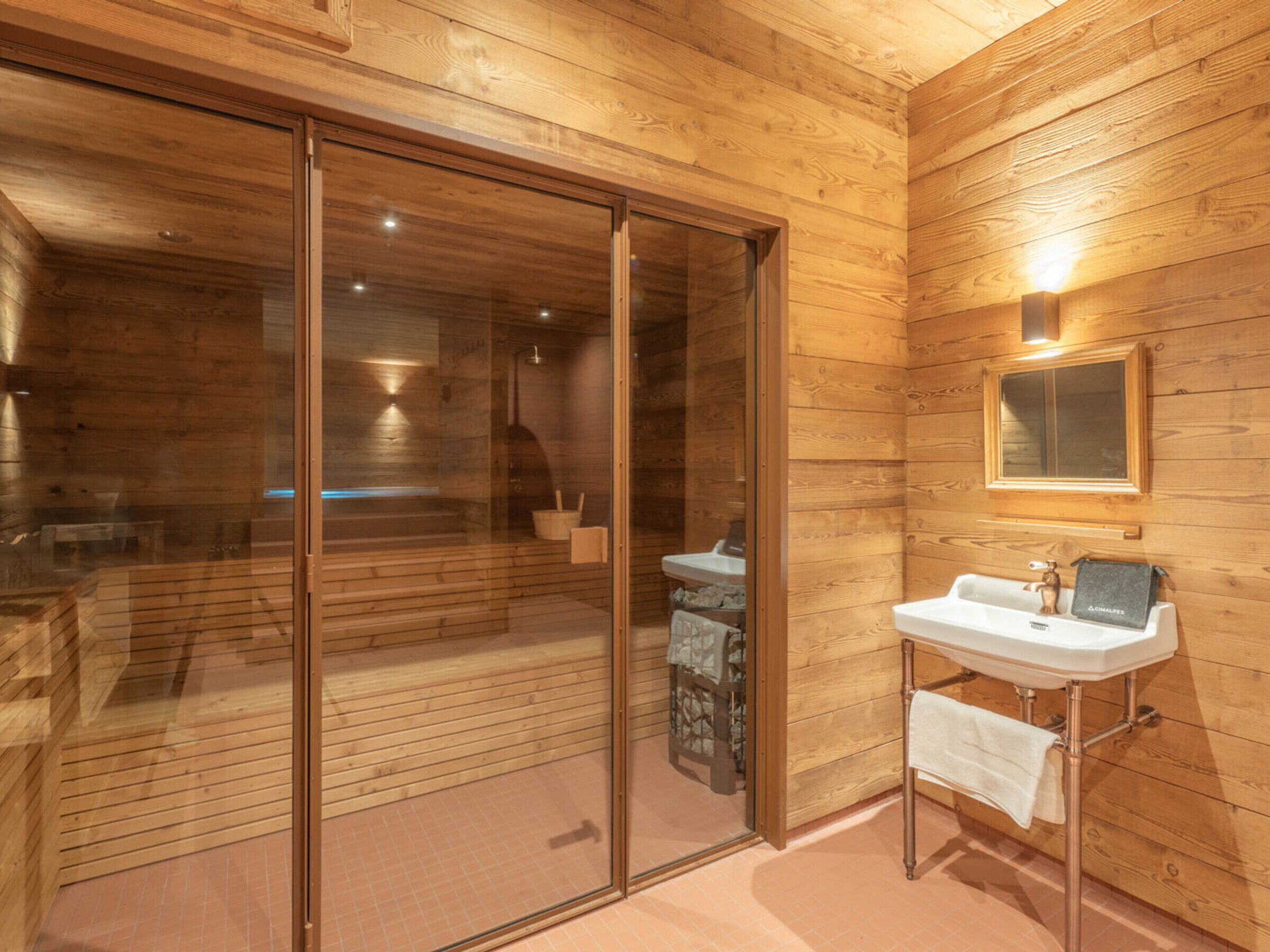 A wooden cabin interior with a glass-enclosed sauna, a sink with a towel, and a mirror on the wall.