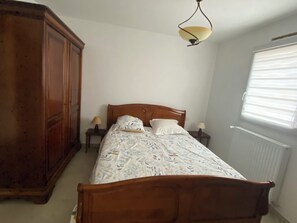 A bedroom with a wooden bed, two bedside tables with lamps, a large wardrobe, and a window with blinds. - La Baie: Family Home for 10 People on the Jade Coast (shared garden) (La Bernerie-en-Retz)