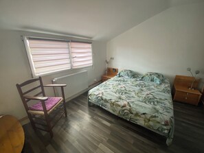 Ein Schlafzimmer mit Bett, Stuhl, Nachttisch, Fenster mit Jalousien und Heizkörper.