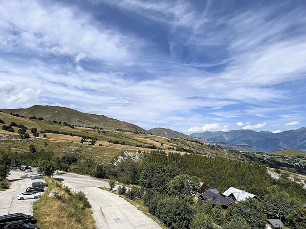 Un paysage rural avec une route, des voitures garées et des maisons entourées de verdure et de montagnes.