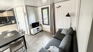 Ein modernes Wohnzimmer mit einem grauen Sofa, einem Fernseher auf einem weißen Schrank, einem Esstisch mit Stühlen und einem durch eine offene Tür sichtbaren Küchenbereich.