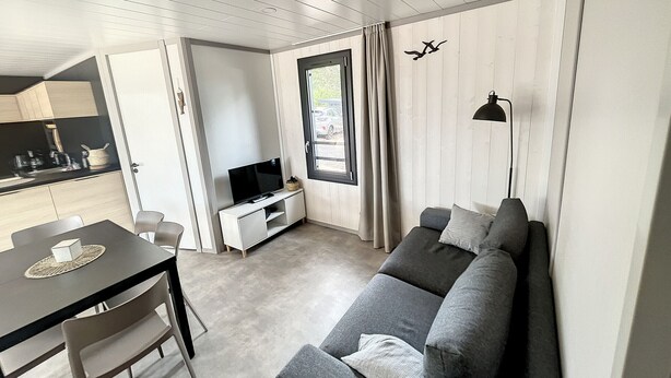 Una sala de estar moderna con un sofá gris, un televisor sobre un mueble blanco, una mesa de comedor con sillas y una cocina visible a través de una puerta abierta.