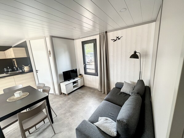 Un salon moderne avec un canapé gris, un téléviseur posé sur un meuble blanc, une table à manger avec des chaises, et un coin cuisine visible à travers une porte ouverte.