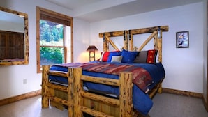 A rustic wooden bed with a headboard, a window with blinds, a lamp, and a framed picture on the wall.