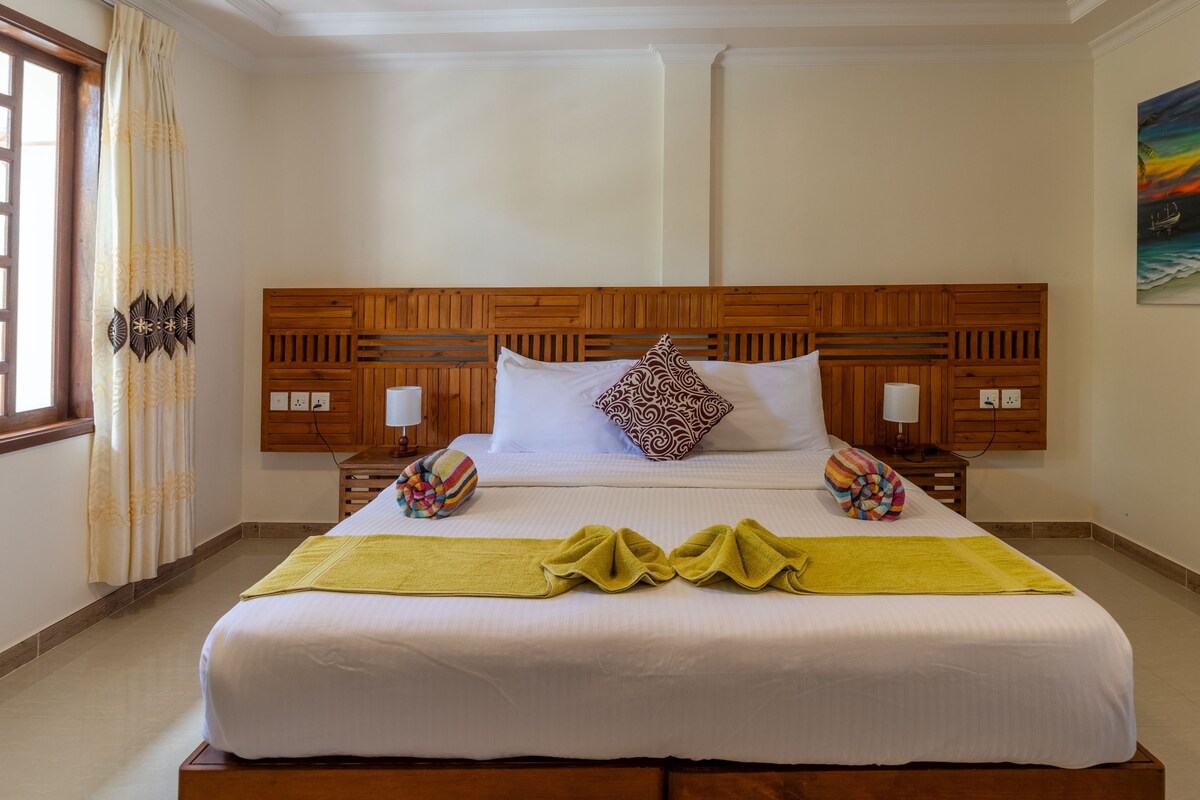 Una habitación de hotel con cabecera de madera, dos mesitas de noche con lámparas, una cama con ropa de cama blanca y una toalla colorida sobre la cama.