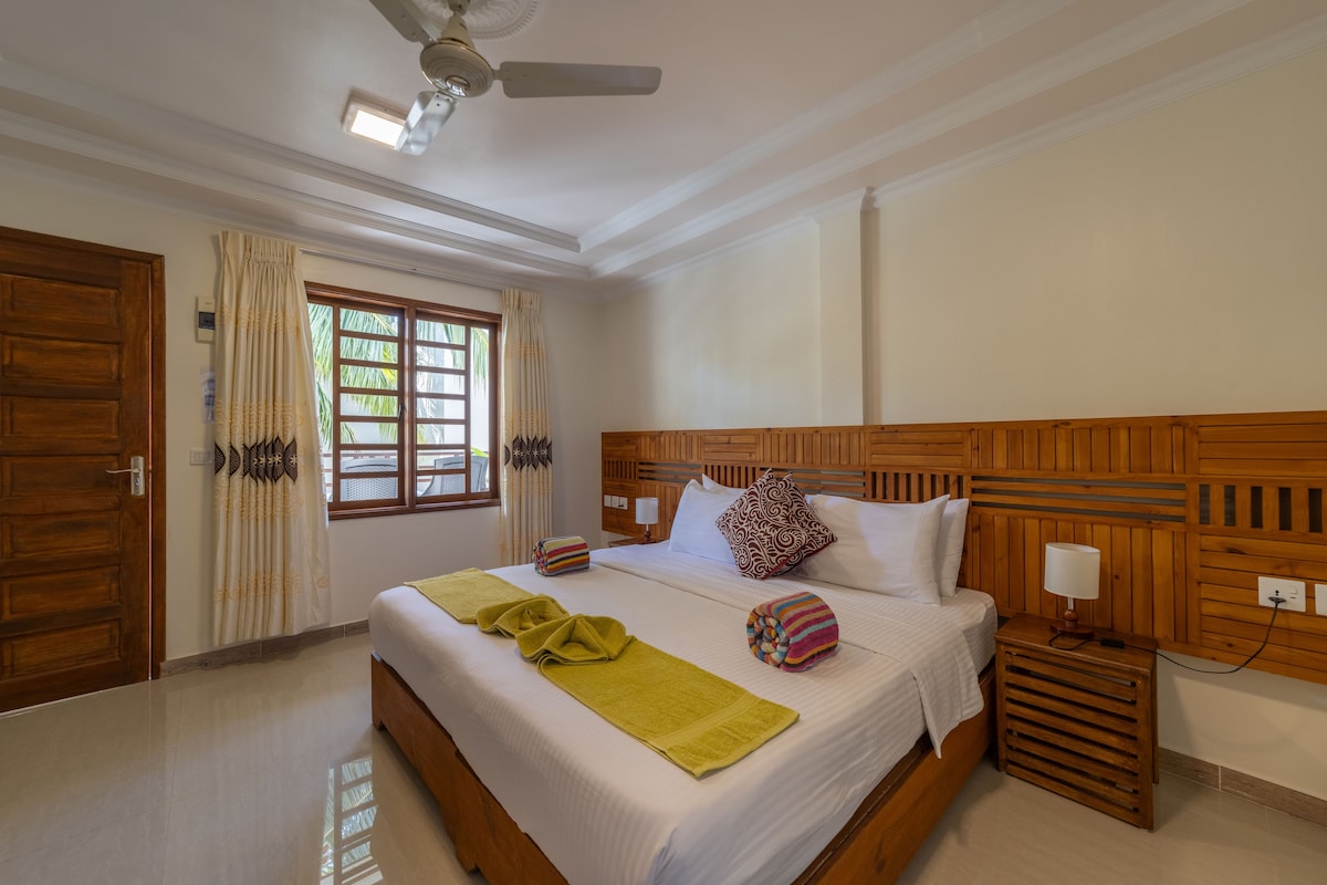 A hotel room with a wooden headboard, a bed with white linens, a bedside table with a lamp, a window with curtains, and a ceiling fan.