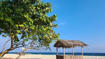 A beach with a thatched structure and a tree providing shade.