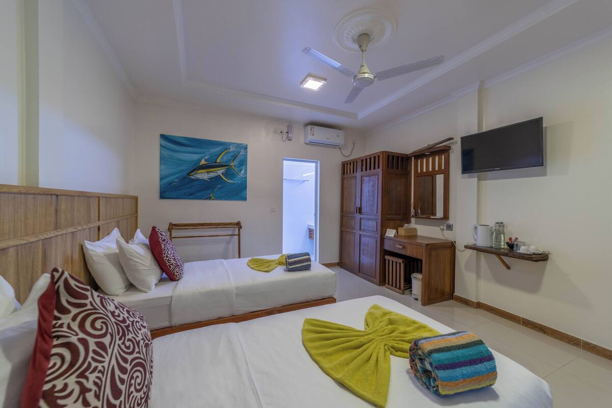 A hotel room with two beds, a ceiling fan, a TV, and a painting on the wall.