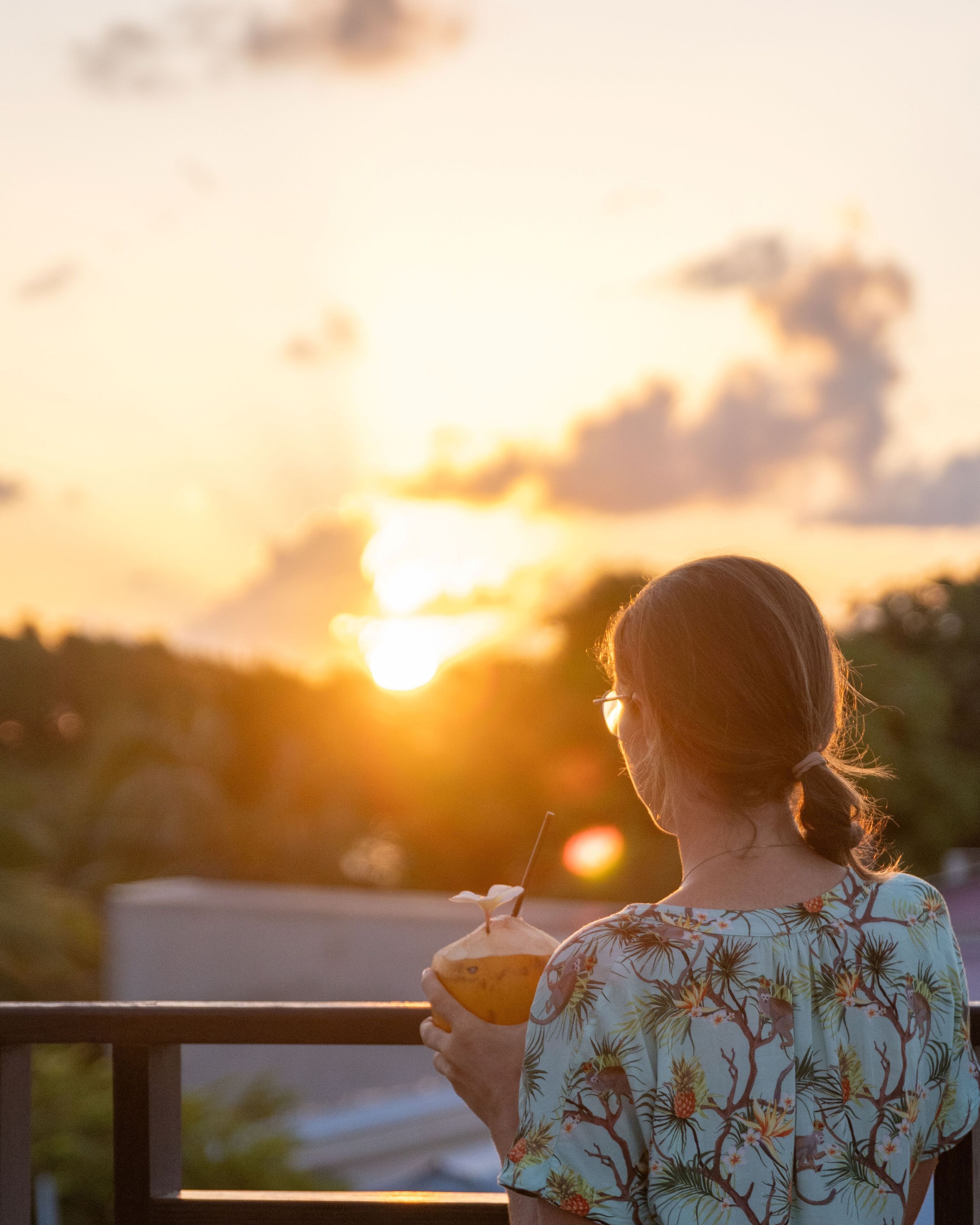 A person holding a drink, overlooking a sunset with a railing in the foreground.