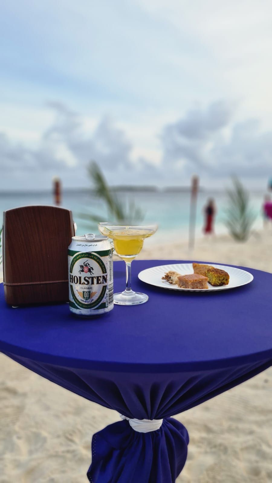 A table with a beer can, a glass of beer, and a plate of food on a beach with a view of the sea.