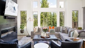 Ein geräumiges Wohnzimmer mit Kamin, großen Fenstern mit Blick auf Bäume, einer Couch mit vielen Kissen und einem Couchtisch mit Dekoration.