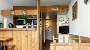 Cocina compacta con armarios de madera, un microondas y un lavabo. Hay una pequeña zona de comedor con una mesa y sillas.