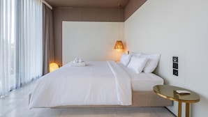 A modern hotel room with a large bed, a bedside table, a lamp, and a view of the outdoors.