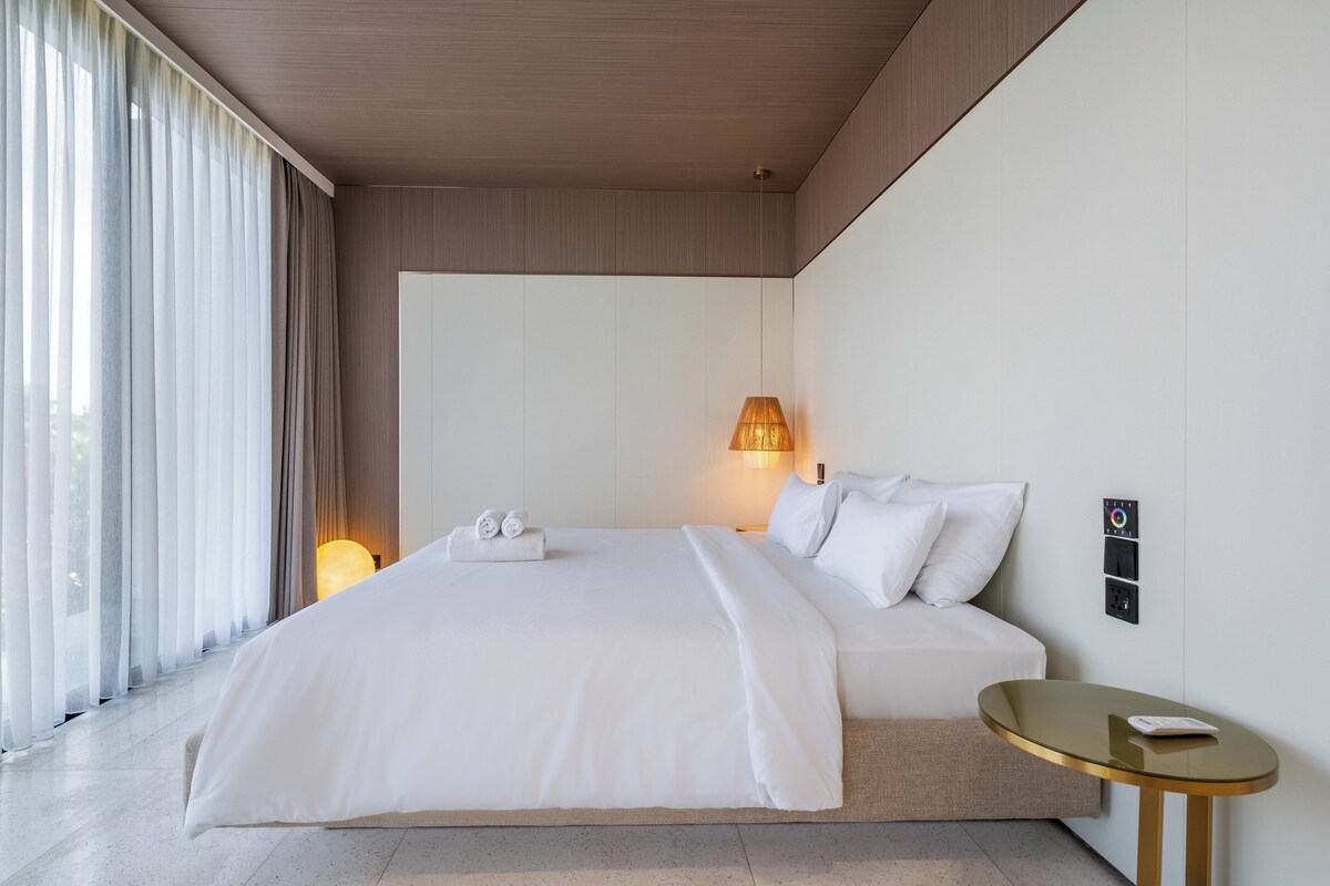 A modern hotel room with a large bed, a bedside table, a lamp, and a view of the outdoors.
