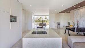 A modern kitchen with a central island, a dining area with a table and chairs, and a view of palm trees through the open space.