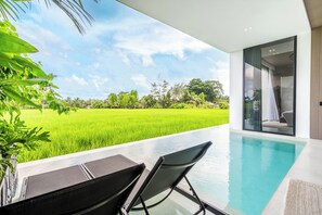 A modern outdoor pool area with a view of a lush green field, a dining table with chairs, and a glass door leading to a room. - Green Flow Villa 8 (Ubud)