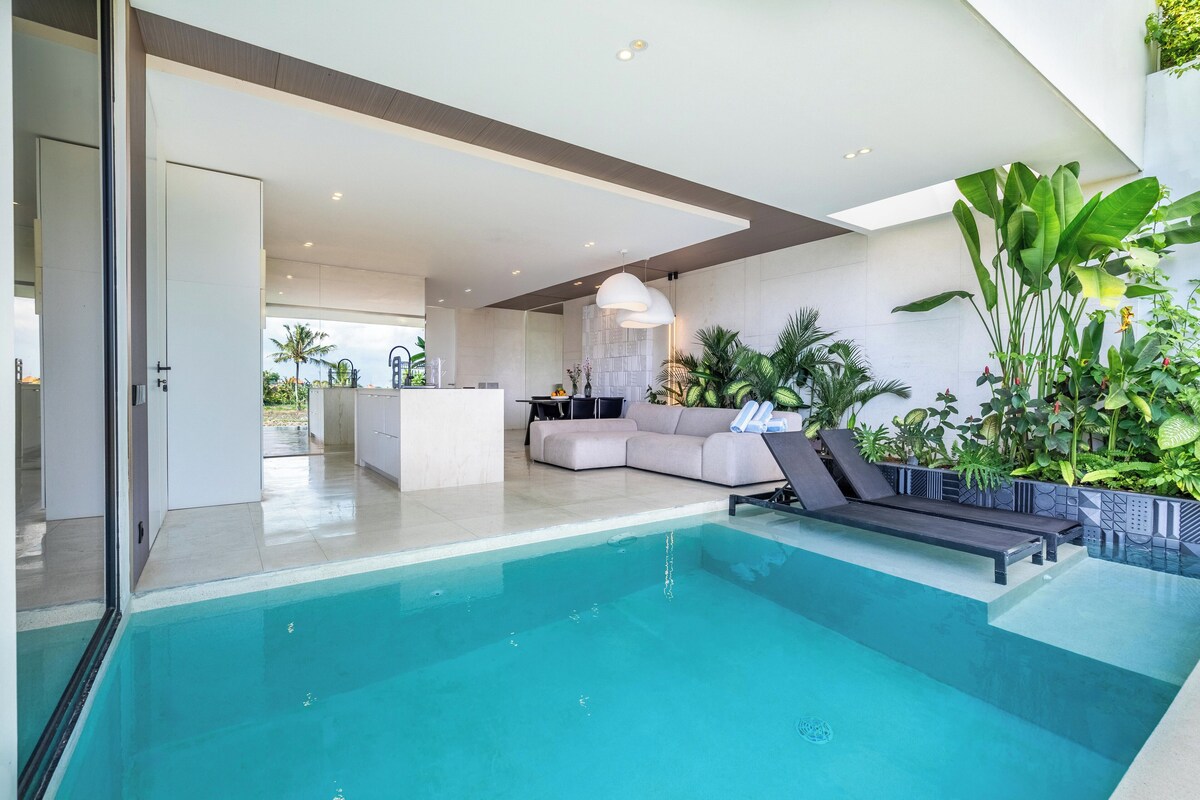 A modern indoor pool area with a lounge chair, a sofa, and a dining area in the background.