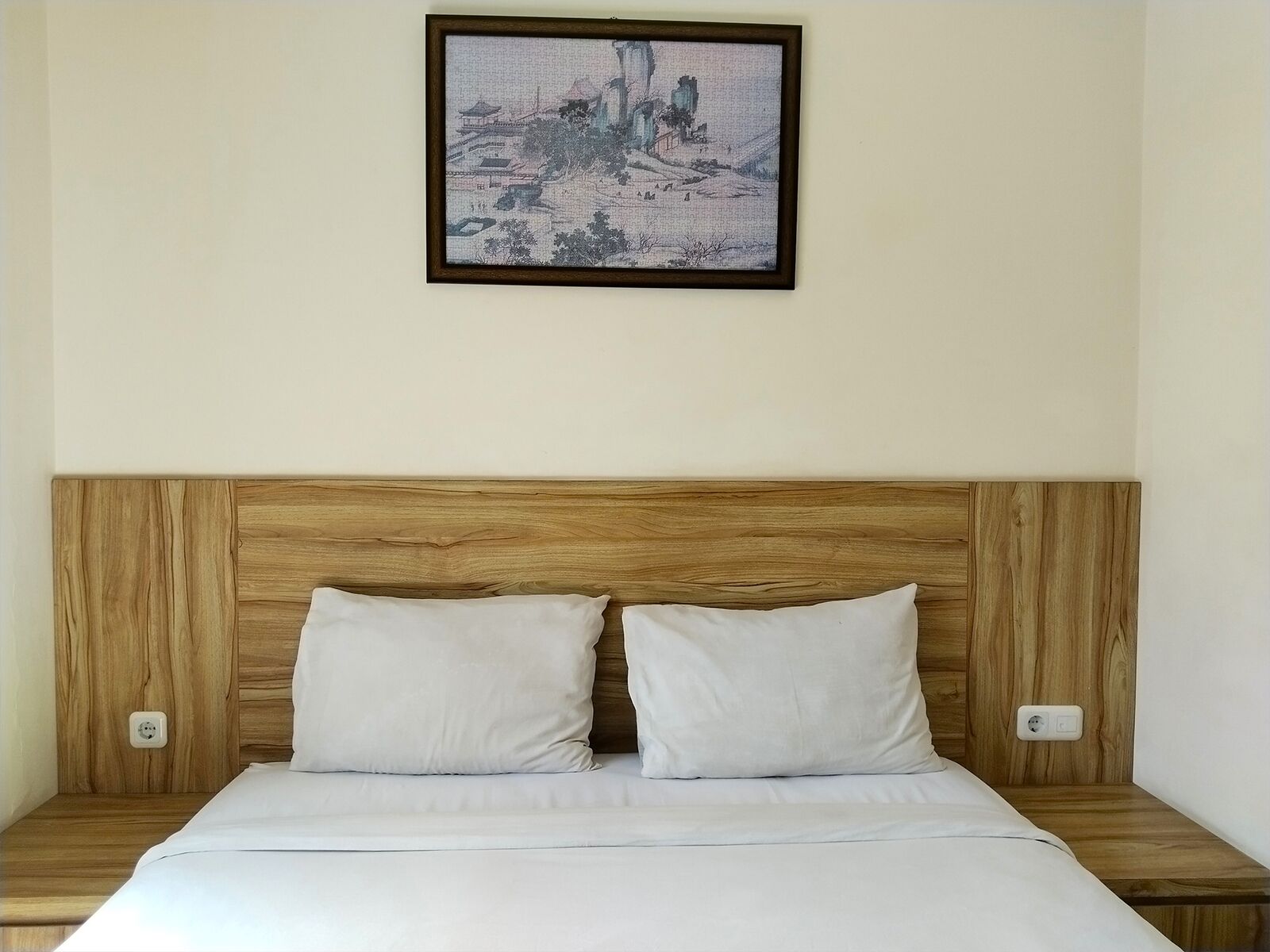 A neatly made bed with a wooden headboard, two white pillows, and a framed artwork above it.