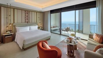 A hotel room with a large bed, a sofa, a dining table, and a balcony with a view of the sea.