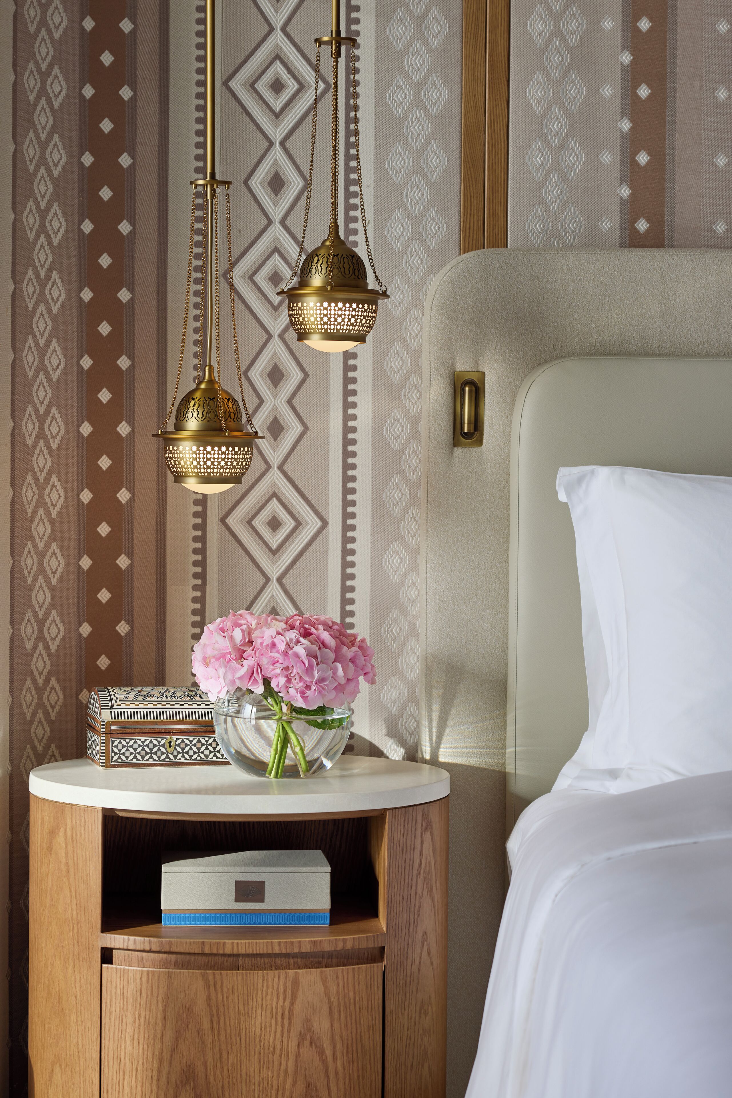 A neatly arranged hotel room with a bed, a bedside table with a lamp, a vase with flowers, and patterned wallpaper.